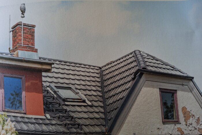 Tegeltak med takfönster, röd tegelsten skorsten och väderkvarn ovanpå, samt en fasad med avskavd puts under en molnig himmel.