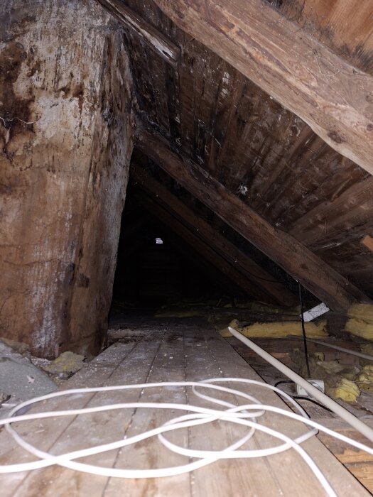 Vind med brädgolv, synliga takbjälkar och isolering. En kabel ligger på golvet.