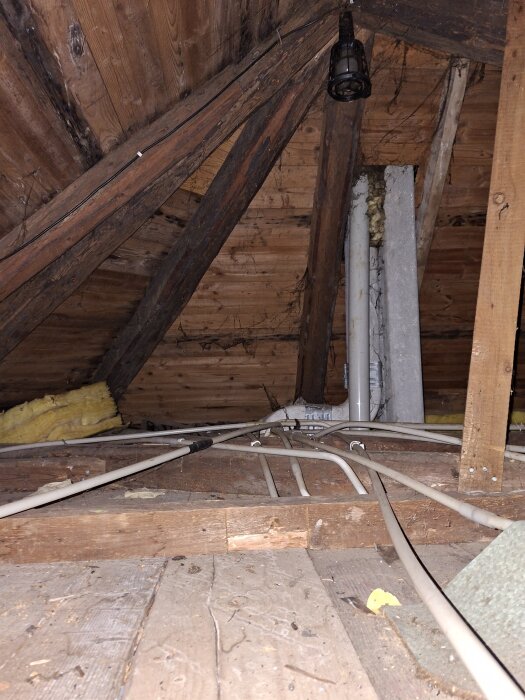 Gammalt tak med träbjälkar på vind, synliga rör och isoleringsmaterial.