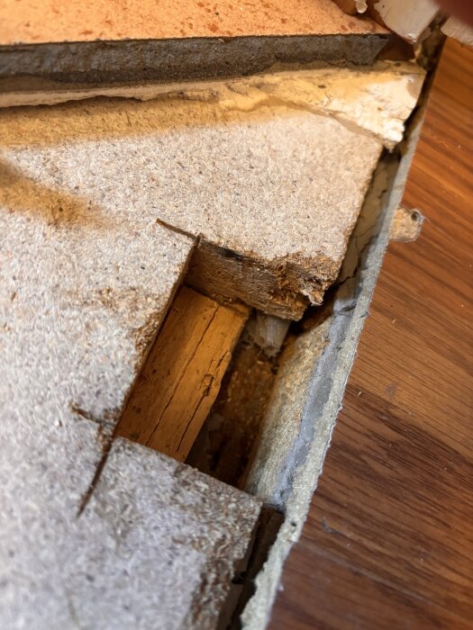 Närbild av ett exponerat golvlag med olika skikt, inklusive gips och spånskiva, ovanpå en träbjälke.
