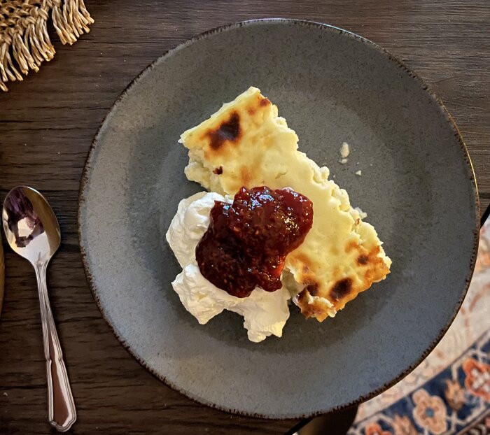 Ostkaka serverad med en klick grädde och hallonsylt på en grå tallrik, fotograferad uppifrån. En sked ligger bredvid tallriken.