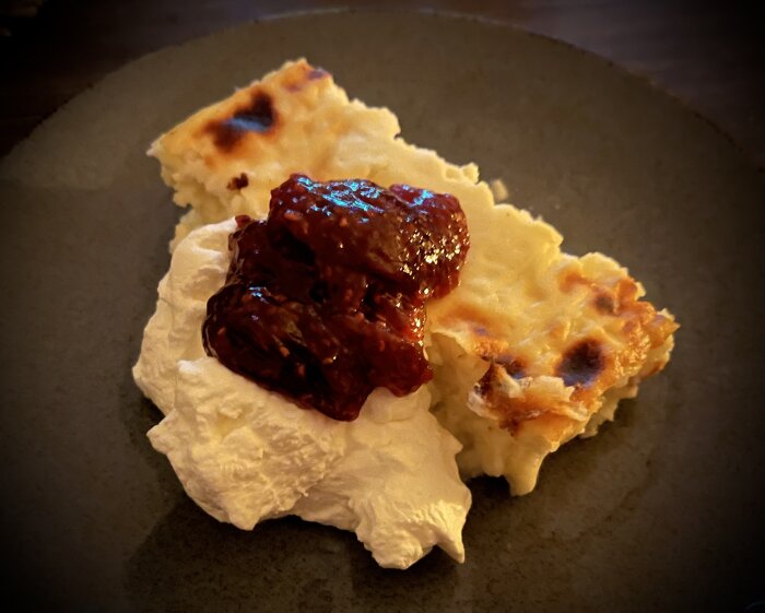 Ostkaka serverad med en klick grädde och hallonsylt på en tallrik.