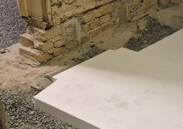 Cellplastskivor ligger mot en ojämn murad vägg, omgärdat av grus.