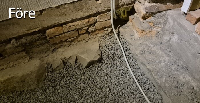 Ojämn murstensvägg med grus lager på marken och ett synligt vitt rör, före installation av cellplast för gjutning av betongplatta.