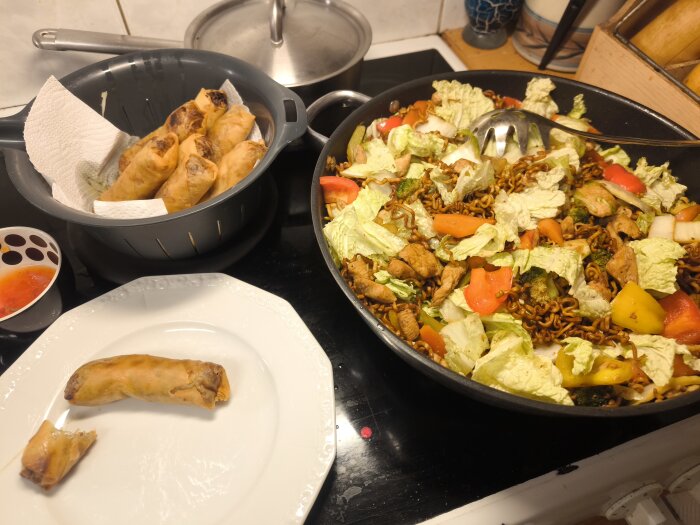 En tallrik med en vårrulle, en panna med nudlar och grönsaker, en sil med fler vårrullar samt en skål med dippsås på en spis.