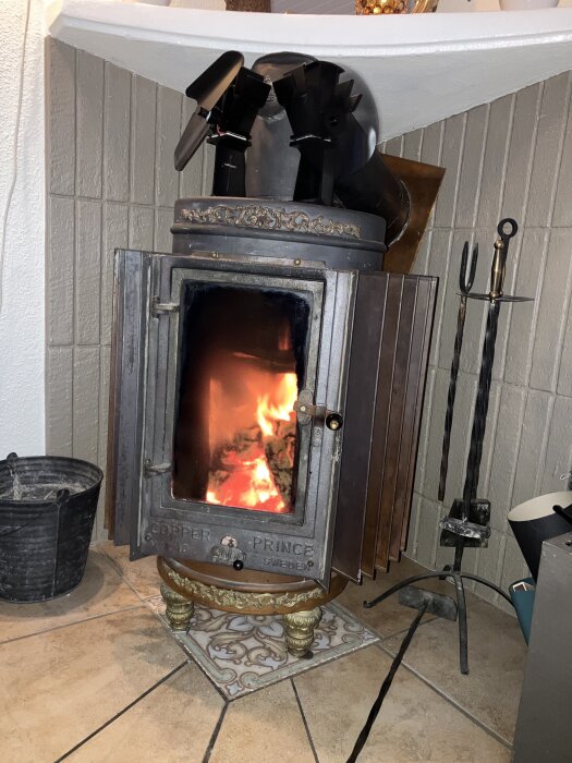 Vedeldad kamin märkt "Cooper Prince" med öppen lucka och synlig eld. Kaminverktyg och askhink står bredvid.