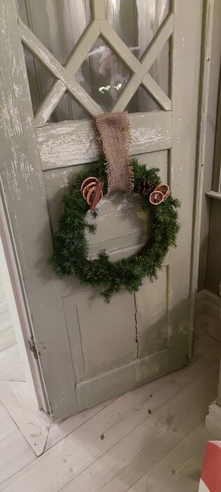 Grön krans med kanel och torkade apelsinskivor hänger på en gammal grågrön trädörr med glasrutor.