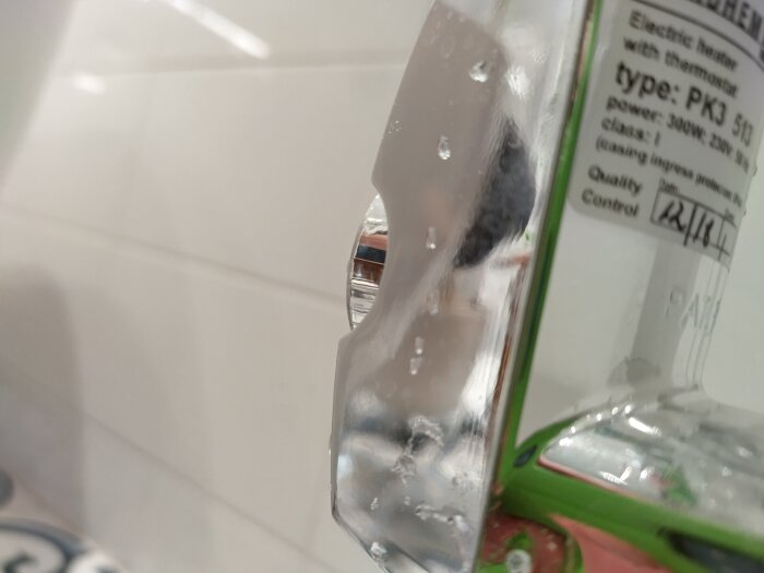 Närbild på en elektrisk handdukstork med en plastplatta, etikett synlig med text, och vattenstänk.