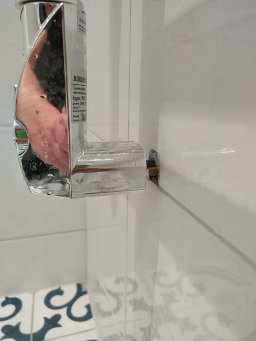 Elektrisk handdukstork monterad på väggen med synliga kablar utan isolering, vattenstänk på torken och kakelvägg i bakgrunden.