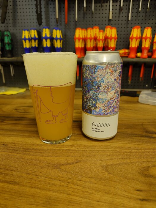 Ölglas och ölburk märkt "Gamma" på en träbänk i ett garage med verktyg i bakgrunden.