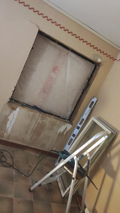 Ett borttaget fönster ersatt med isolering i en vägg. Framför syns ett avvägningsverktyg och en stege med en borrmaskin.