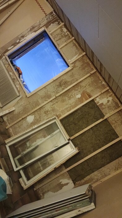 Vägg med borttagna fönster och exponerad isolering påbörjad renovering, med verktyg och byggmaterial synligt i bild.