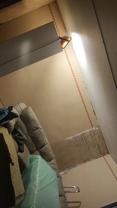 Inuti ett rum under renovering med en lampa på väggen, synlig isolering och byggmaterial. Plast och glasfiberväv är framträdande.