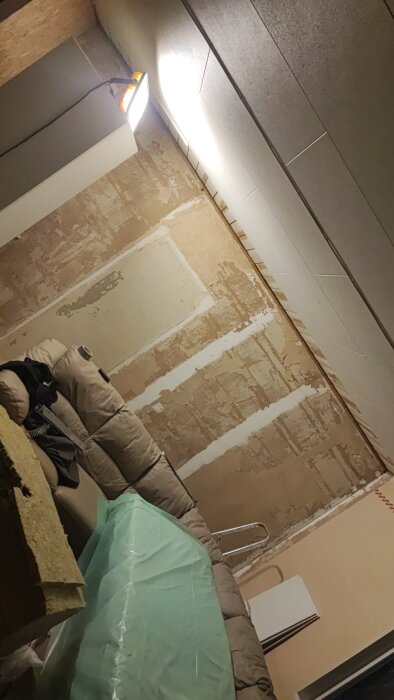 Rum med rivna väggar och tak, belyst med bygglampa; soffa, byggmaterial och verktyg är också synliga.