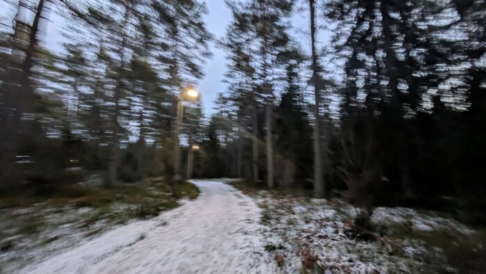 En oskarp bild av en snötäckt skogsstig med gatulampor som lyser upp vägen i skymningen, omgiven av höga träd.