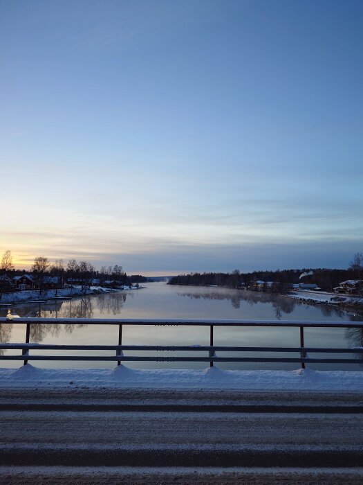 Snöig väg och bro med utsikt över stilla flod, omgiven av träd och hus i soluppgång eller solnedgång, med klar himmel ovanför.