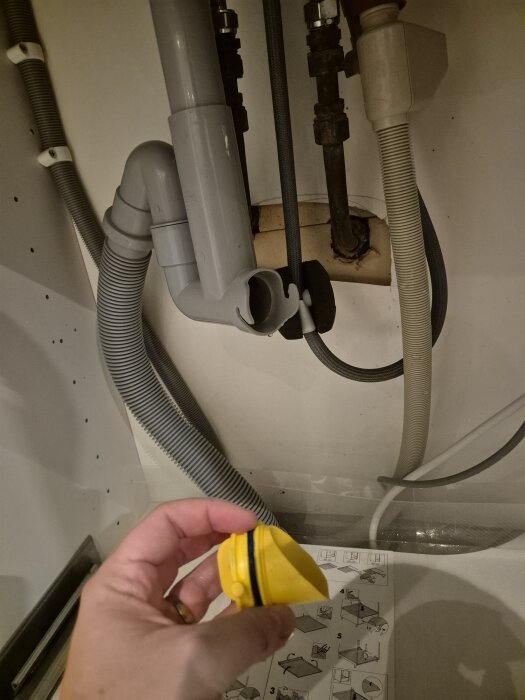 En hand håller i en gul plastdel framför rör och slangar under ett handfat, med instruktioner synliga i bakgrunden.