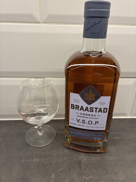Flaska Braastad VSOP cognac och ett tomt cognacsglas på en grå bänkskiva med vit kakelbakgrund.