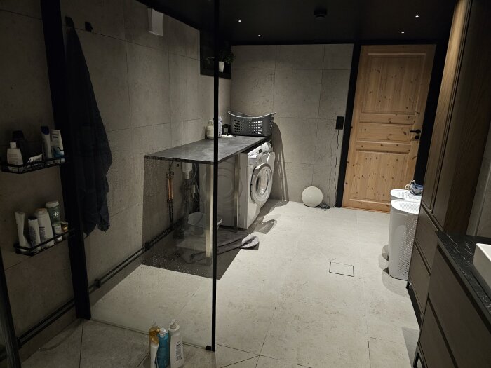 Tvättstuga med svart tak, grå väggar och golv, tvättmaskin under hylla, dörr i trä, hyllor med flaskor och korgar för tvätt.