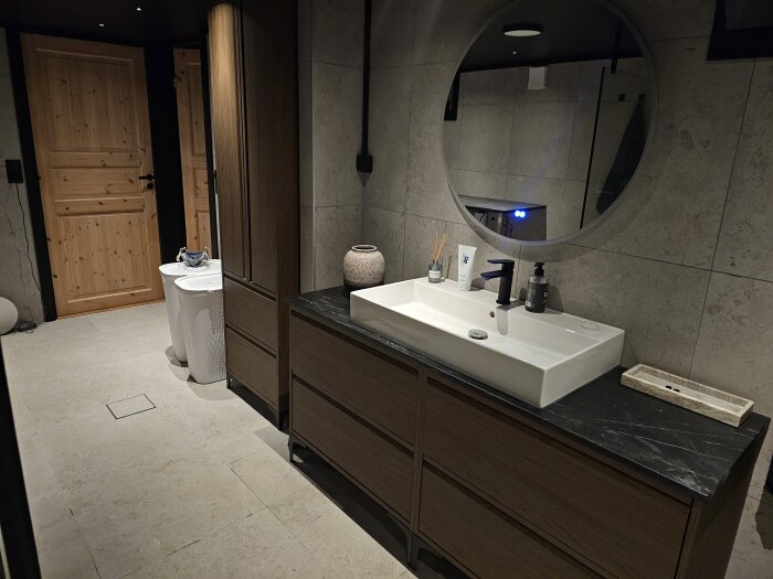 Modernt badrum med trädetaljer, stor spegel och svart tak. Vitt tvättställ på svart bänkskiva. Dörr av ljust trä och två vita behållare.