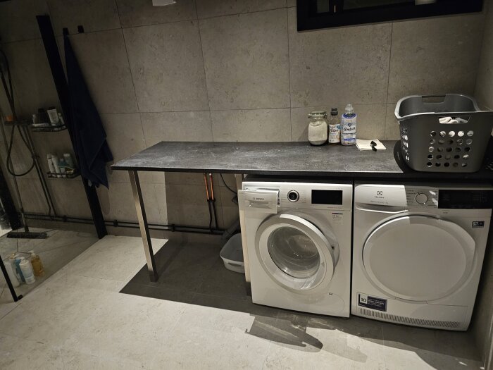 Tvättstuga med vit tvättmaskin och torktumlare under en svart bänkskiva. Grå väggar och golv, korg och tvättmedel på bänken.