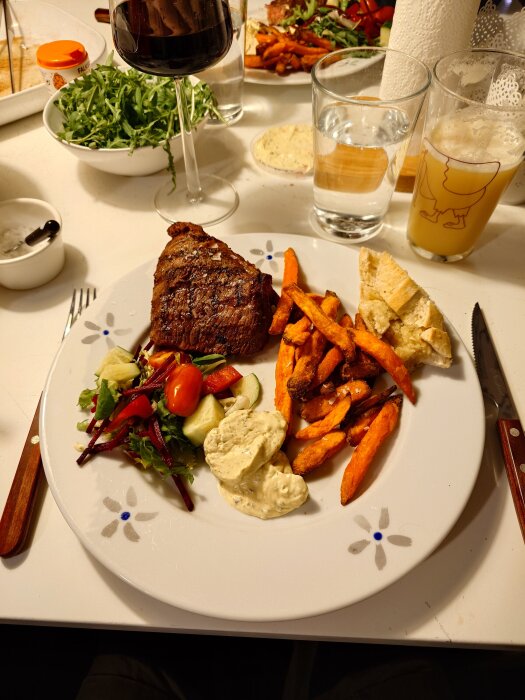 Grillad rostbiffservering med sötpotatis pommes, sallad och sÄs pÄ en tallrik. Vin, dryck och sallad i bakgrunden.