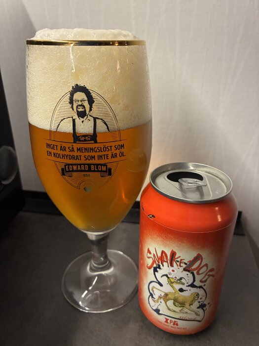 Ett glas med öl från Edward Blom's märke bredvid en burk Snake Dog IPA mot en vit bakgrund.