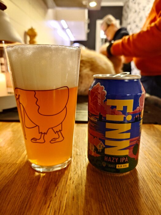 Ett glas skummande hazy IPA och en färgglad ölburk på ett träbord, personer och en katt syns i bakgrunden.