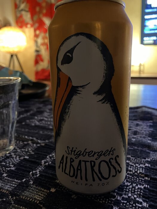 Ölburk med etikett föreställande en albatross och texten "Stigbergets Albatross NEIPA 7.0%" på ett bord med röd och blå bakgrund.