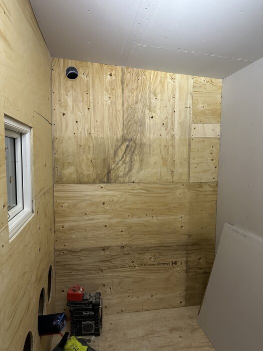 Rum under renovering med plywoodväggar, ett fönster och några verktyg. Svarta fläckar syns på en vägg, och material är ordnat på golvet.