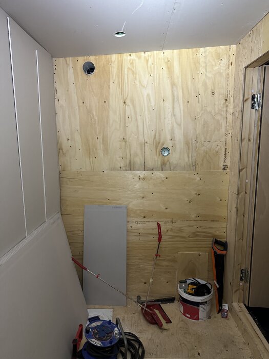 Byggmaterial och verktyg i ett garage, med plywood och gipsskivor vid väggen, samt olika verktyg som målarpensel och såg på golvet.