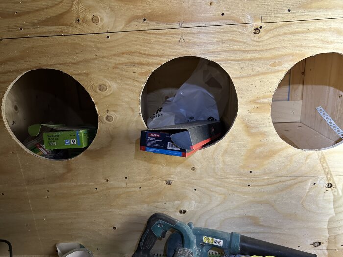 Tre runda hål i plywoodpå en vägg med verktyg och förpackningar insatta. Verktyg och byggmaterial syns i bilden.