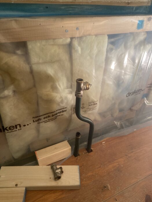 Rör nära väggen över en träbotten, inklädda i ångspärr och isolering. Träbitar och rörkopplingar ligger på golvet.