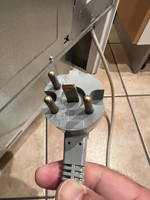 Grått eluttag med fyra stift för anslutning av spis, hålls i handen, visar detalj av köksgolv och apparatens baksida i bakgrunden.