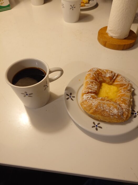 Kaffekopp bredvid ett wienerbröd på en dekorativ tallrik, uppdukat på ett matbord.