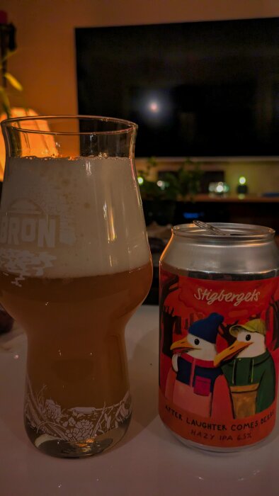 Ett glas med skummande öl bredvid en burk Stigbergets Hazy IPA, 6,5%, i ett upplyst rum med dekorationer.