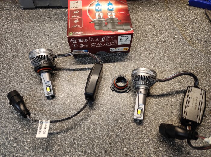 Två Luxtar Hero LED-bilglödlampor med kablar och lösa socklar ligger på ett grått bänkskiva, med en röd kartong i bakgrunden.