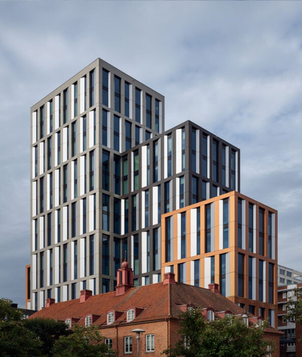 Höga, moderna byggnader i vitt och orange reser sig bakom ett äldre, rött tegeltak, omgivna av gröna träd under en molnig himmel.