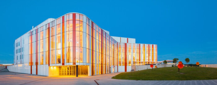 Byggnad med glasfasad i vitt och orange, med mjuka hörn mot en blå himmel. Gräsmatta med runda konstverk i förgrunden.