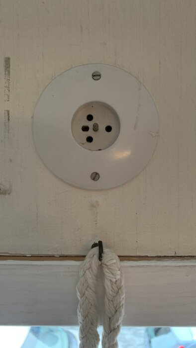 Gammalt eluttag ovanför fönster, installerat på 70-talet, med flera hål för kontaktpinnar. Stort runt väggfäste med skruvar.