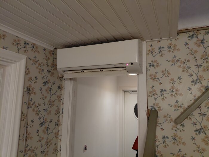 Väggmonterad luftvärmepump inomhusenhet i ett rum med blommig tapet och träpanel i taket, begränsad plats under taket.