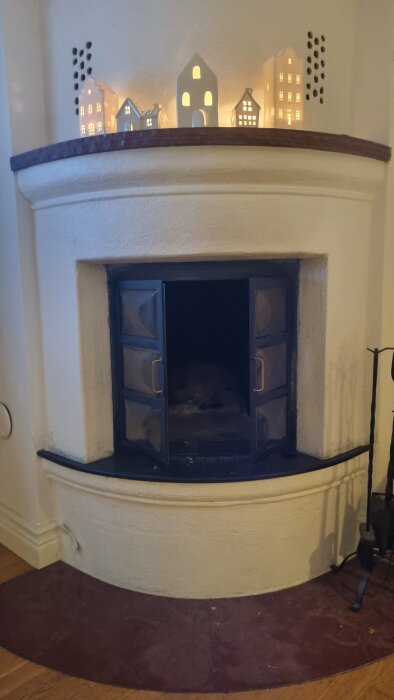 Vit eldstad med öppna dörrar och dekorativa hus på toppen, i ett hus med självdrag.