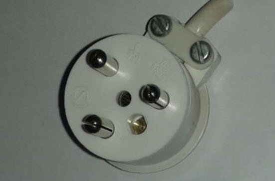 Jordad kronstickpropp med fyra stift och vit plast, liggande på en ljus yta. Koppling för äldre elektriska lampor eller uttag.