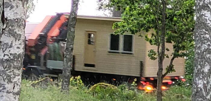 Friggebod transporteras med kranbil genom skogsområde, sedd mellan träd.
