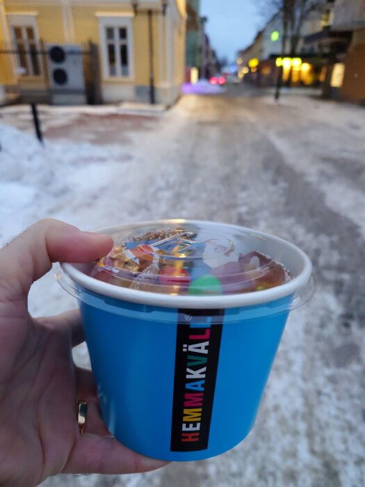 En hand håller en blå burk med godis från Hemmakväll framför en vintergata med snö och hus i bakgrunden.