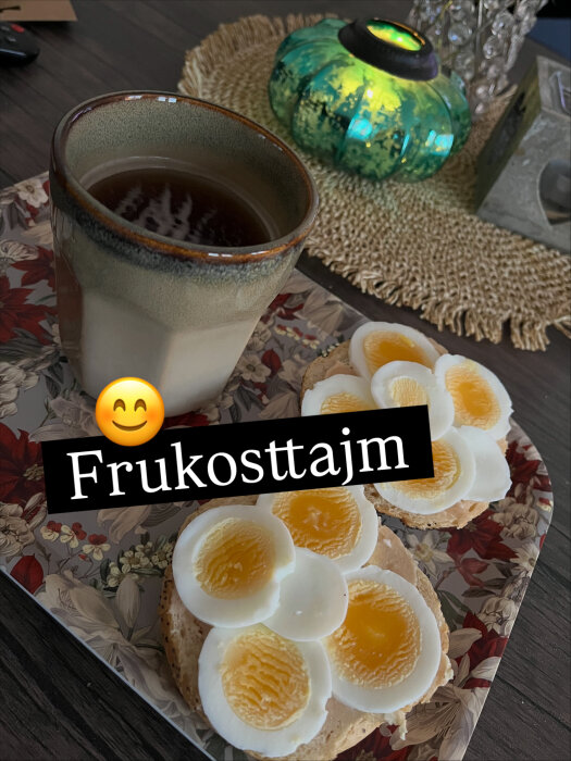 Kaffekopp och mackor med skivat ägg på ett blommigt underlägg, bredvid ett tänt ljus i en färgad ljushållare.