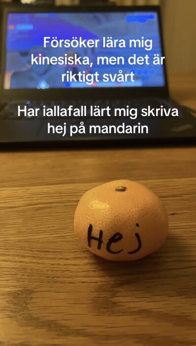 Mandarin på ett bord med ordet "Hej" skrivet på den, en dator i bakgrunden visar text om att lära sig kinesiska.