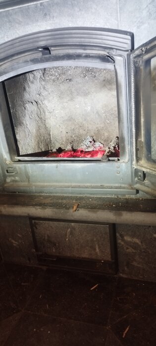 Öppen eldstad med glödande kol i en kamin med öppen glasdörr, omgiven av sotig interiör och grå stenyta.
