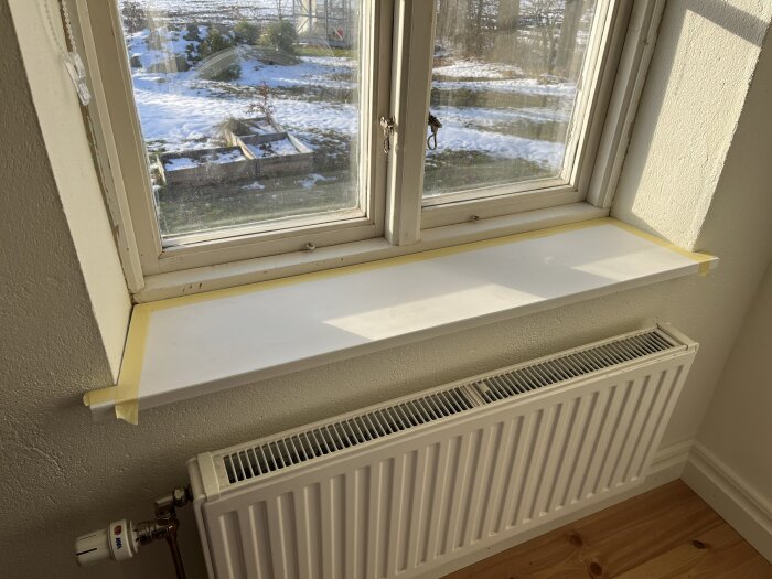 Fönsterbräda med maskerande tejp på kanterna, klart för silikonering. Utsikten visar snötäckt trädgård och en kallvinterdag.