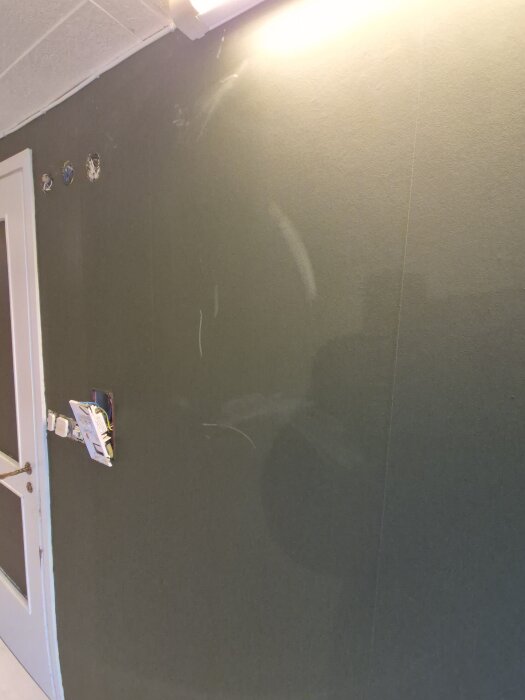 Grönmålad vägg med vita märken som liknar gipsdamm, nära dörr och uttag, under lampa.